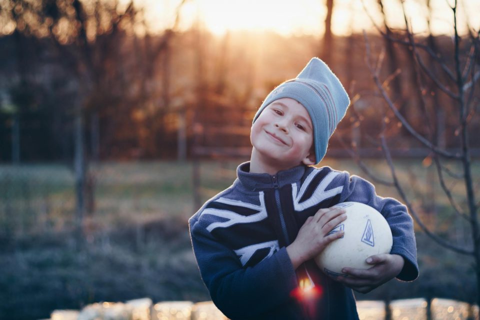 selective focus photography of boy wearing blue united kingdom print zip up jacket carrying white ball