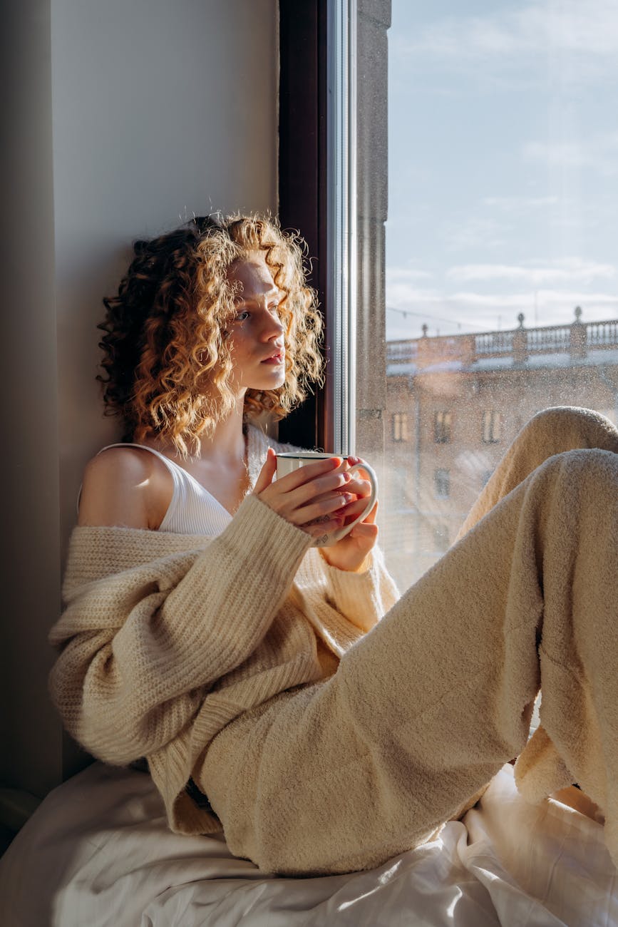 woman in white tank top and brown cardigan sitting by the window