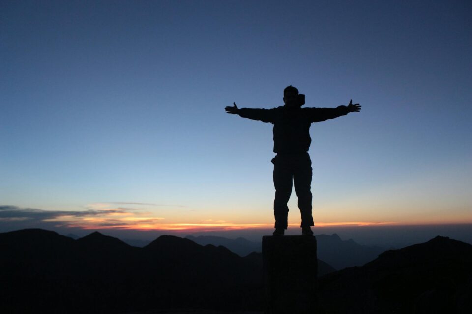anonymous male climber standing on mountain top with outstretched arms at sundown