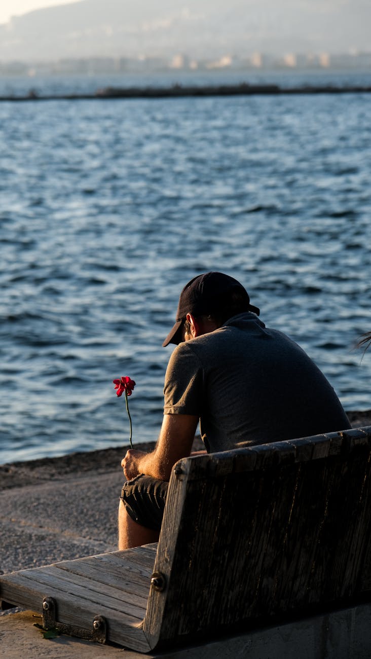 a man holding a flower sitting alone on a bench by the sea